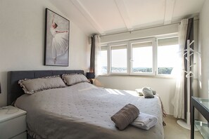 Un dormitorio con cama, mesita de noche, ventana con cortinas y un cuadro en la pared.