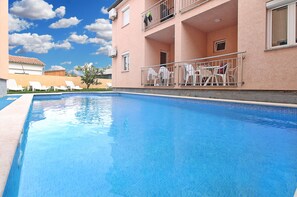 Une piscine à l’eau d’un bleu cristallin, entourée de chaises longues blanches et d’un bâtiment rose doté de balcons.