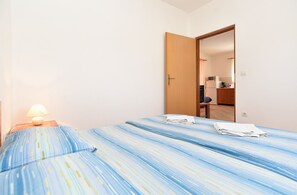 Une petite pièce simple, avec un lit, une table de chevet dotée d’une lampe et une porte donnant sur un coin cuisine.