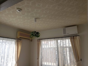 Una habitación con techo estampado, un equipo de aire acondicionado, una planta colgante y un ventanal con cortinas.