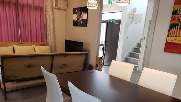 A modern living room with a dark wooden table, white chairs, a wooden sofa with cushions, a flat-screen TV, and a decorative wall piece. - Resortstay in American village (Chatan)