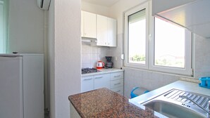 Une cuisine de taille réduite avec des meubles blancs, une cuisinière à gaz, une machine à café et une fenêtre qui laisse entrer la lumière naturelle.