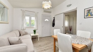 Een woonkamer met een bank, een eettafel met stoelen, een klein bijzettafeltje met een plant, een witte deur en een raam met gordijnen.