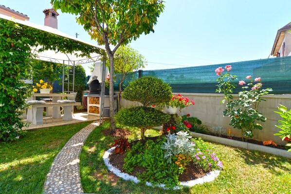 Relax in a serene garden setting surrounded by lush plants and flowers, a stone bench, and an outdoor oven.