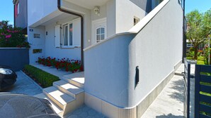Een modern gebouw met een witte gevel, een trap die naar een veranda leidt, en een kleine tuin met rode bloemen.