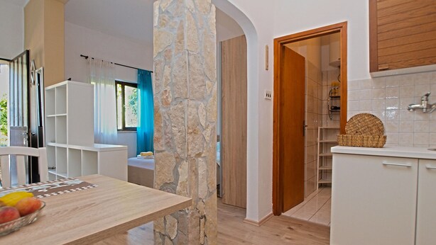 A modern kitchen with white cabinetry, a stone accent wall, and a wooden dining table with fruit. - Studio apartment 1364/12839 (Istria - Pula), 2500m from the beach (Pula)