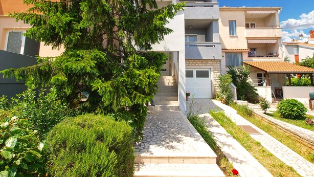 Un immeuble résidentiel moderne à deux étages, avec un chemin de pierre menant à l’entrée, entouré d’une végétation luxuriante et surplombé d’un ciel bleu clair.