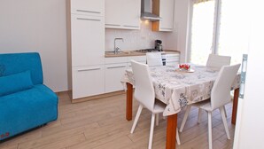 Une cuisine compacte avec des meubles blancs, une cuisinière et un évier. On y trouve une table à manger avec des chaises, ainsi qu’un canapé bleu dans l’espace salon.