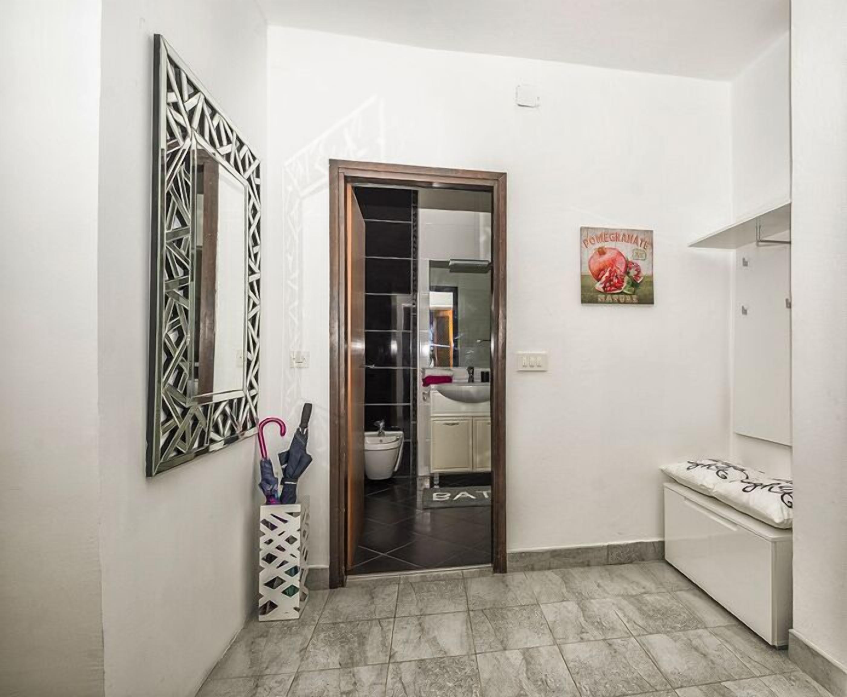 A bathroom with a white tiled floor, a pomegranate-themed wall decoration, a mirror with a geometric frame, and a white bench with a cushion.