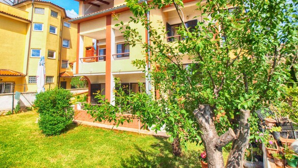 A multi-story building with balconies, a garden area with trees and shrubs, and a clear blue sky.