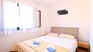 A neatly made bed with two towels, a wooden headboard, a bedside table, and a window with sheer curtains.