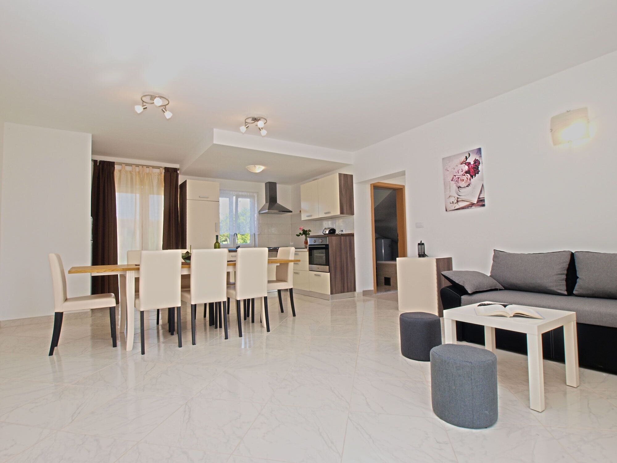 A modern living room with a sofa, ottomans, a dining table with chairs, and a kitchen in the background.