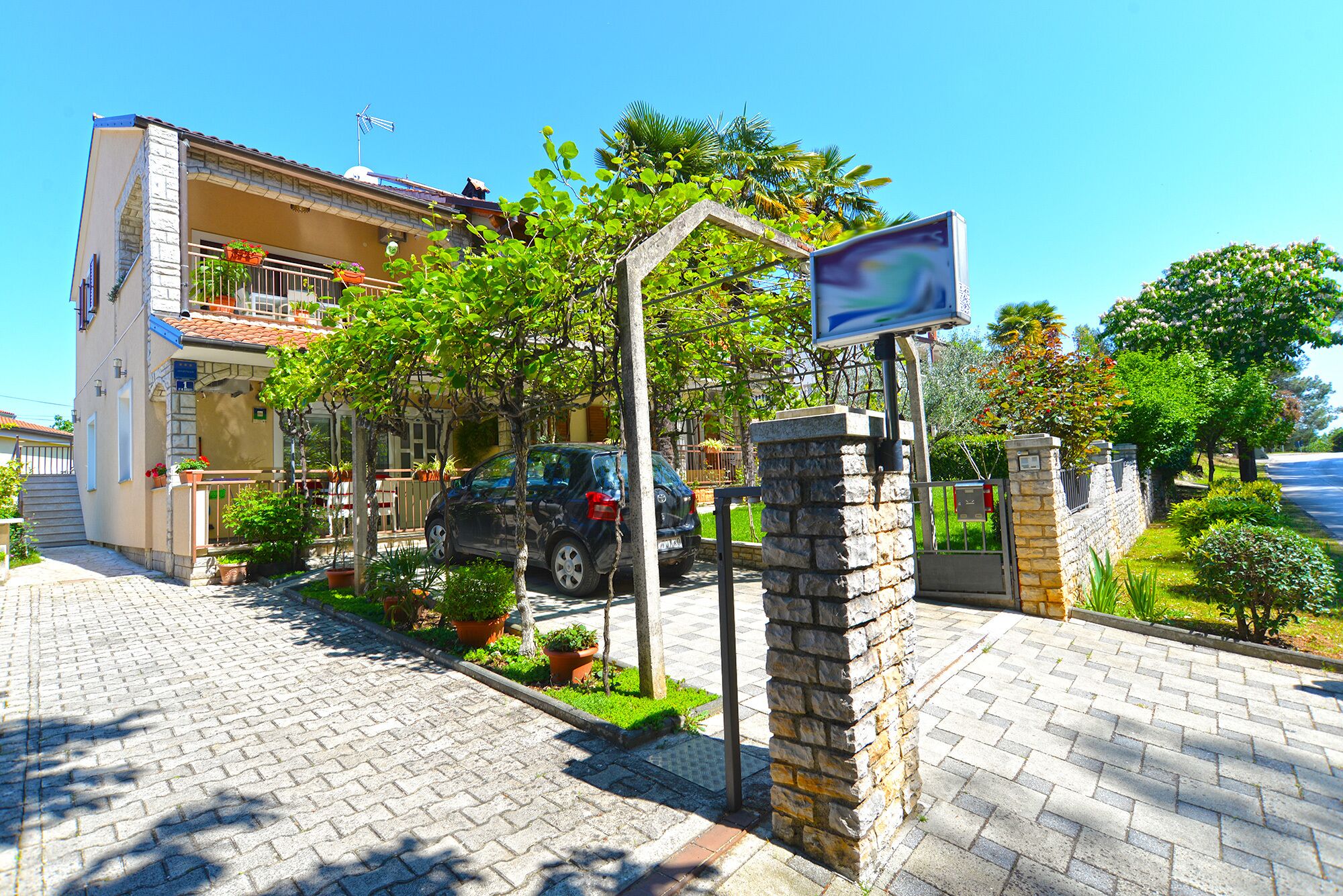 A residential building with a paved driveway, a parked car, and a stone wall with a sign.