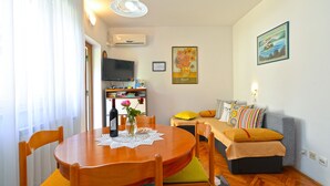 A cozy living room with a round wooden dining table, a sofa with colorful cushions, a television, and a framed painting on the wall. - Apartment 1377/13010 (Istria - Poreč), 800m from the beach (Poreč)