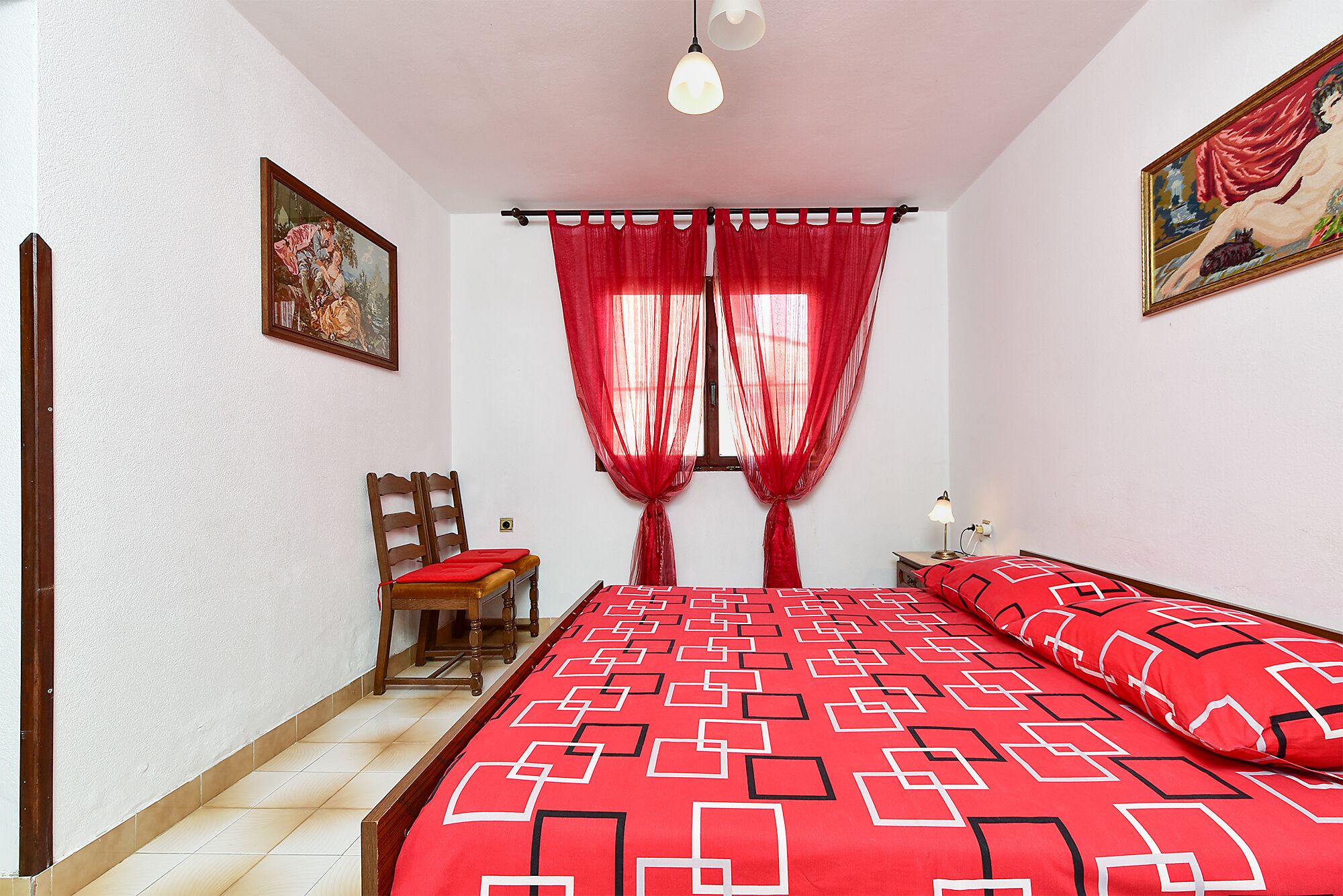 A bedroom with a bed, a chair, a nightstand, a lamp, and two paintings.