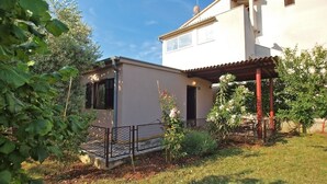 Ein zweistöckiges Haus mit überdachter Terrasse, einem umzäunten Garten und blauem Himmel.