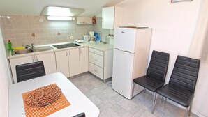 A compact kitchen with a sink, stove, and refrigerator, two black chairs, and a clock on the wall.