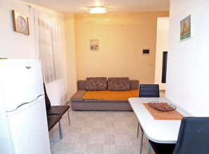 Ett kompakt vardagsrum med ett litet kök, en soffa med mönstrade kuddar, ett matbord och en liten tavla på väggen.
