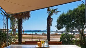 Mesa con sillas bajo una pérgola, con vista a una playa con barcos y cielo despejado.