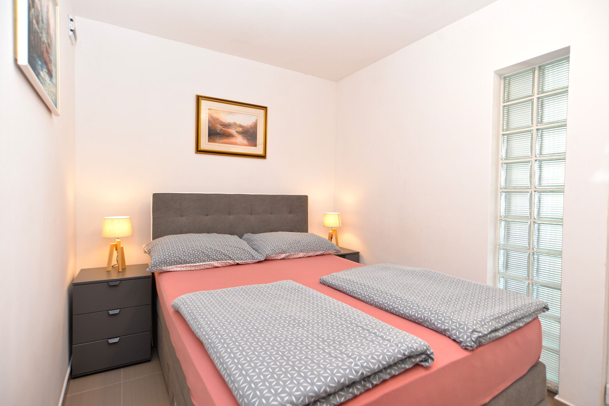 Une chambre à coucher comprenant un lit, deux tables de chevet, une commode, une fenêtre avec des stores et un tableau encadré accroché au mur.
