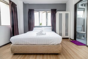 A neatly made bed with a rolled towel, a wooden floor, a purple rug, and a bathroom visible through an open door. - Garden City Melaka Raya by IdealHub (Malacca City)