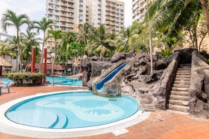 A swimming pool with a rock slide, surrounded by palm trees and a building in the background. - Garden City Melaka Raya by IdealHub (Malacca City)