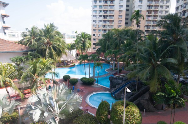 Relax by the resort pool surrounded by lush greenery and palm trees, featuring a curved pool with a central water slide and lounge chairs.