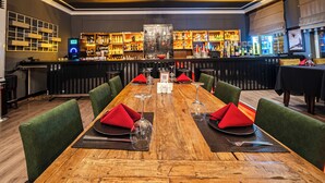 A restaurant with a long wooden table set for dining, green upholstered chairs, and a bar with shelves of bottles and glasses. - Grand Yazıcı Uludağ (Bursa)