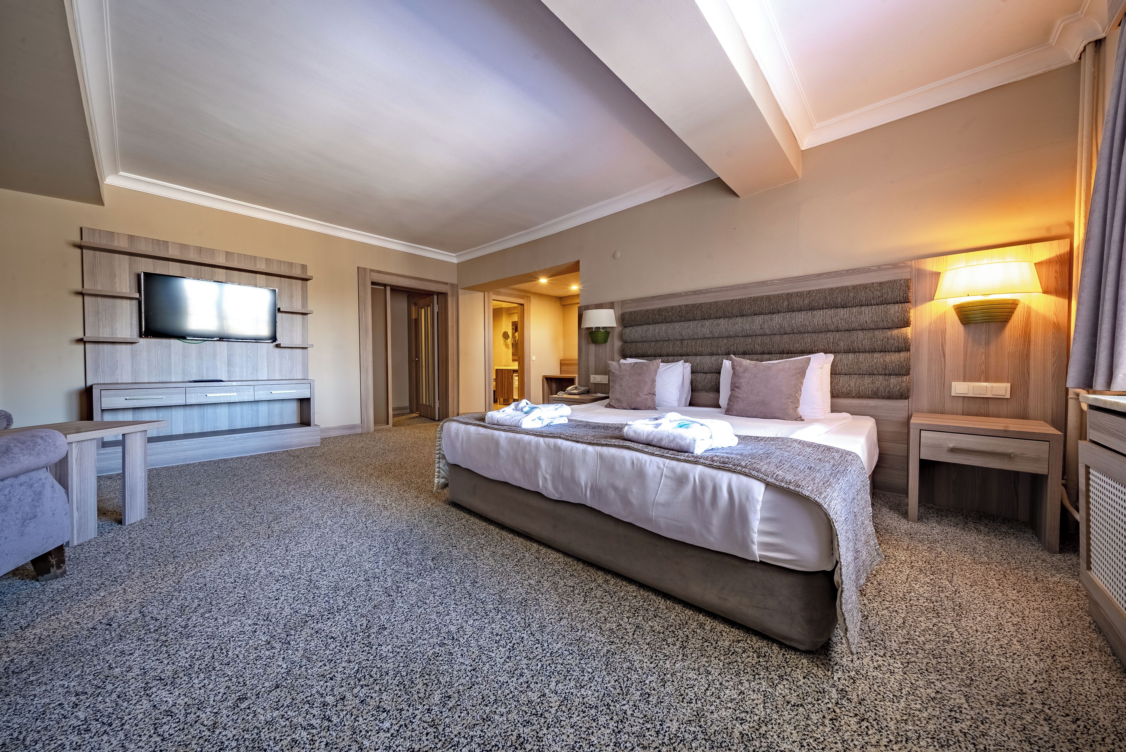 A spacious hotel room with a large bed, a flat-screen TV mounted on a wooden panel, a bedside table, and a lamp.