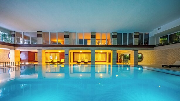 A large indoor swimming pool with a clear view of the pool area, a seating area with tables and chairs, and a staircase leading to an upper level. - Grand Yazıcı Uludağ (Bursa)