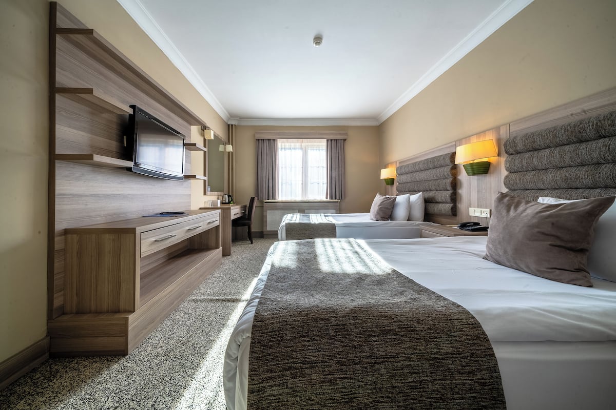A hotel room with a flat-screen TV mounted on a wooden wall, a desk with a chair, two beds with headboards, a window with curtains, and a bedside lamp.