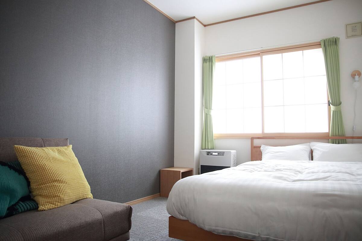 A hotel room with a bed, a small bedside table, a wall-mounted heater, and a window with curtains.
