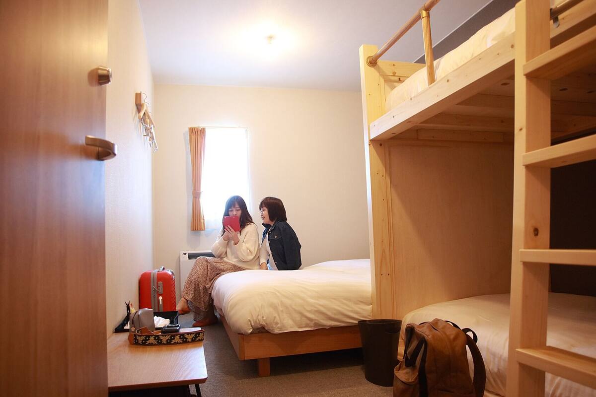 A dormitory room with bunk beds, a desk with a suitcase, and a window with curtains.