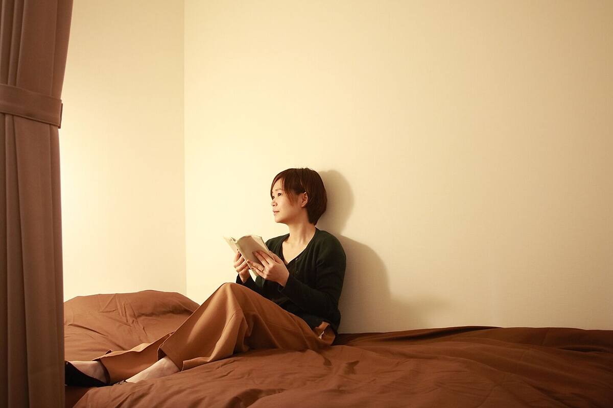 A person sitting on a bed in a room with a plain wall and a window with curtains.