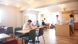 A modern dining area with wooden tables and chairs, a kitchen area with a sink, and a large window providing natural light. - The Stay Wakkanai- Hostel (Wakkanai)