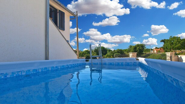 Una piscina con agua azul cristalina, rodeada por un edificio blanco con una ventana y una escalera de metal.