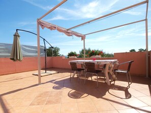 En udendørs patio med et bord og stole, en pergola med et tag, samt plantekasser med planter.