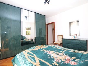 Et soveværelse med stor garderobe, en seng med blomstret sengetæppe, en stol, et kommode og et spejl på væggen.