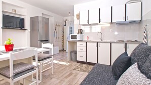Cocina compacta con gabinetes blancos, microondas y estufa. Hay un comedor con mesa y sillas blancas, y un sofá gris en la sala.