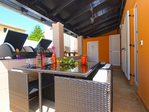 Un patio al aire libre con muebles de mimbre, una mesa con bebidas y flores, y vista al exterior del edificio.