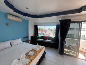 Une chambre d’hôtel avec un grand lit, un petit coin salon avec des coussins, une fenêtre avec des rideaux et un balcon avec une table et des chaises.