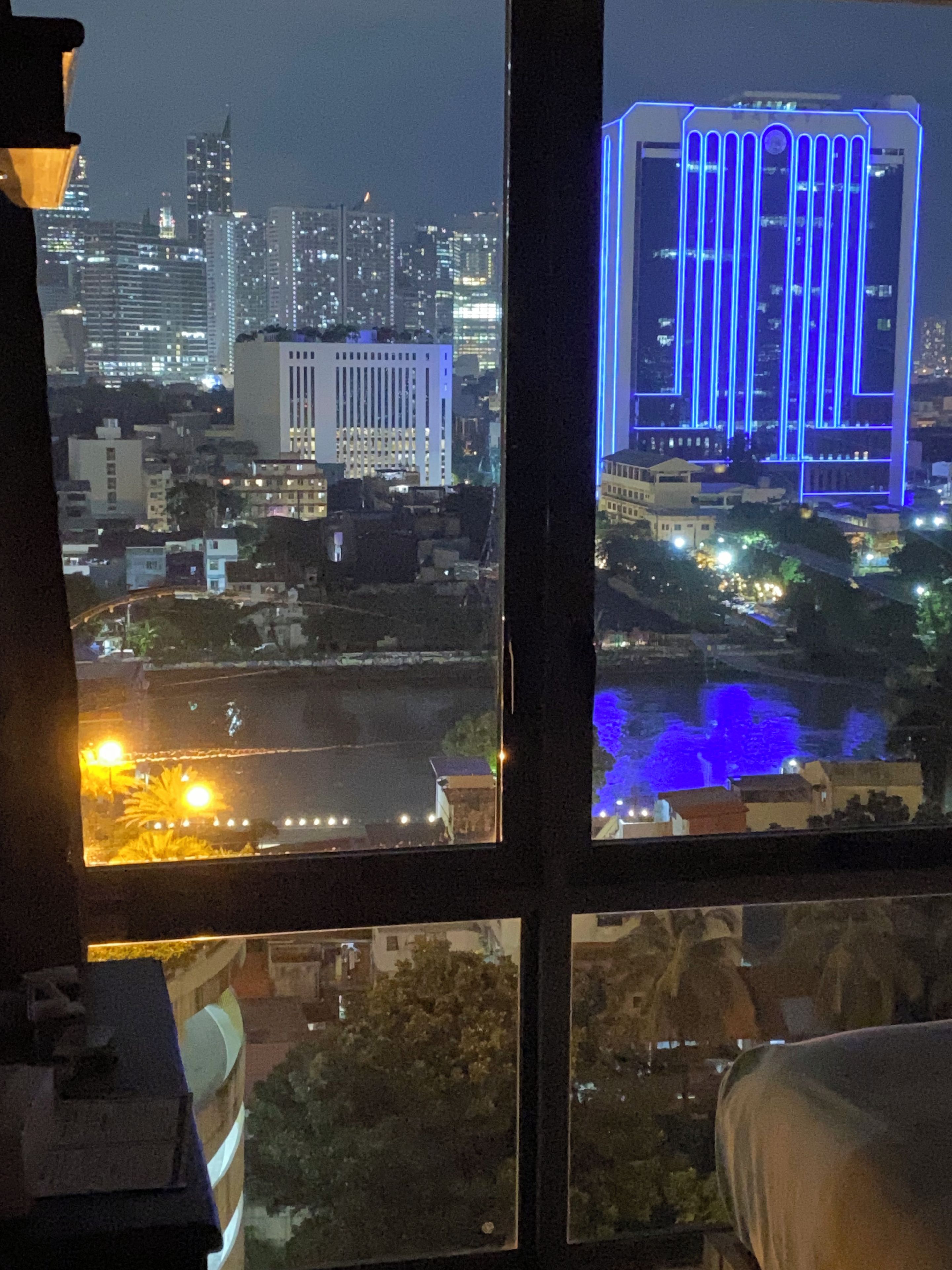 A nighttime cityscape through a window with a view of a river, buildings, and a bridge with blue lighting.