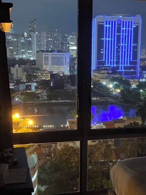 A nighttime cityscape through a window with a view of a river, buildings, and a bridge with blue lighting. - 2 bedroom condo available at Tivoli Garden Residences Luxury Resort makati (Metro Manila)