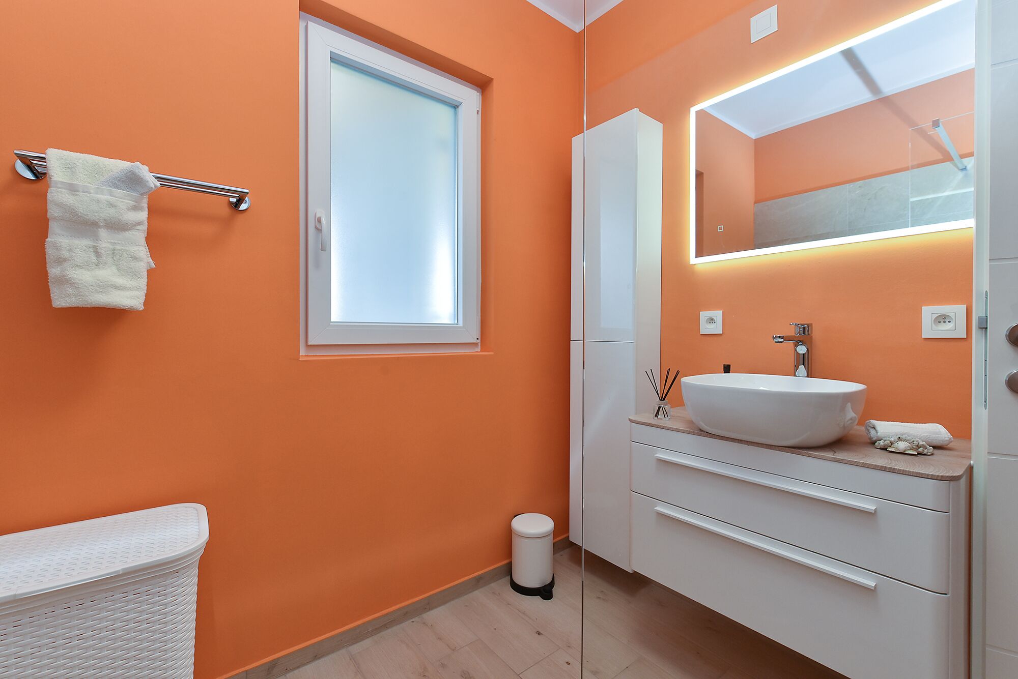 Un baño con una pared naranja, un lavamanos blanco, un espejo y una ventana.