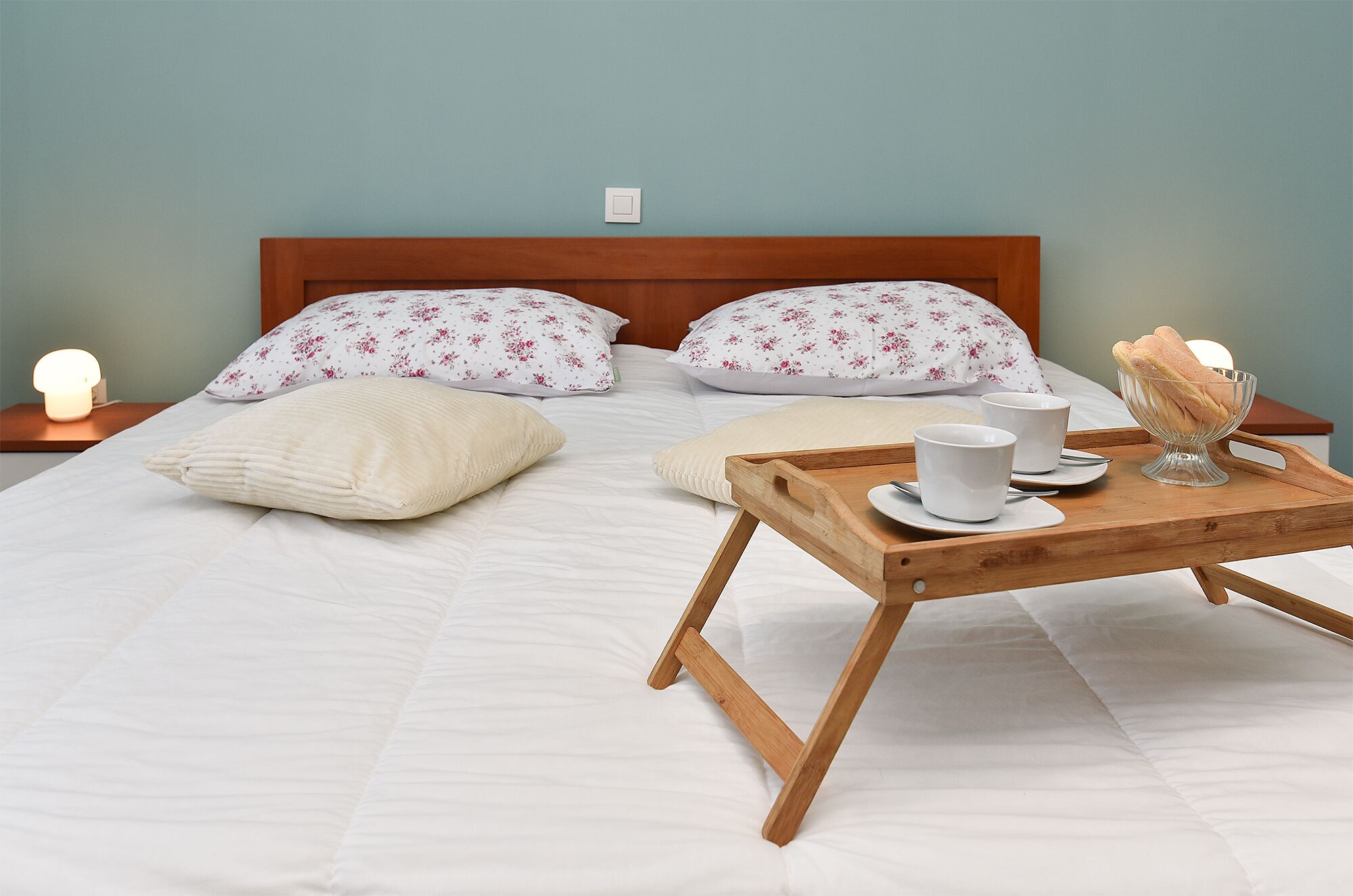 A neatly made bed with white bedding, two floral pillows, a bedside table with a lamp, and a tray with a cup and dessert.