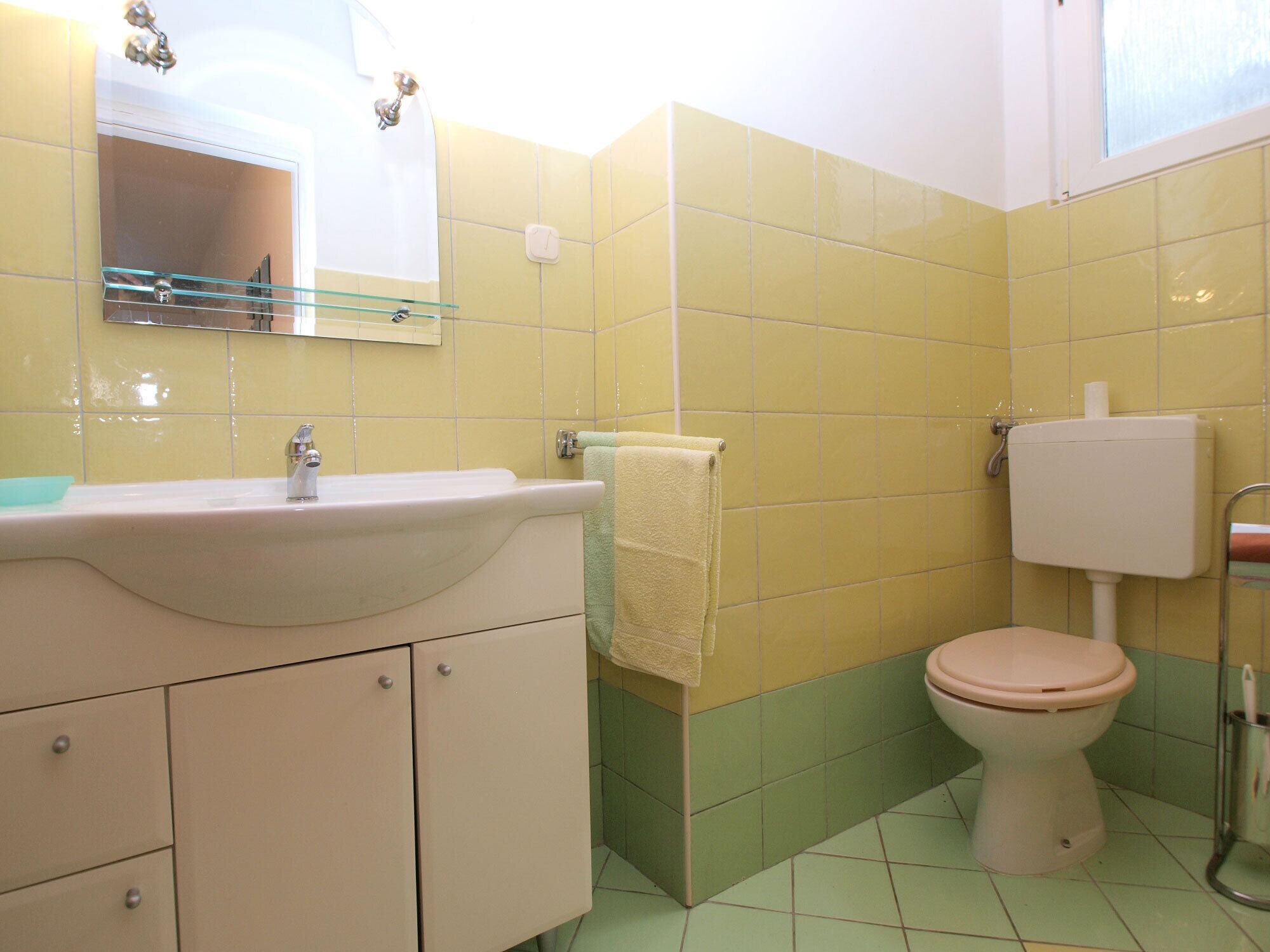 A bathroom with yellow and green tiles, a white sink, a mirror, and a toilet.