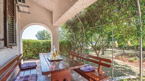 An outdoor patio with a wooden table and benches, surrounded by greenery and a clear view of the sky.