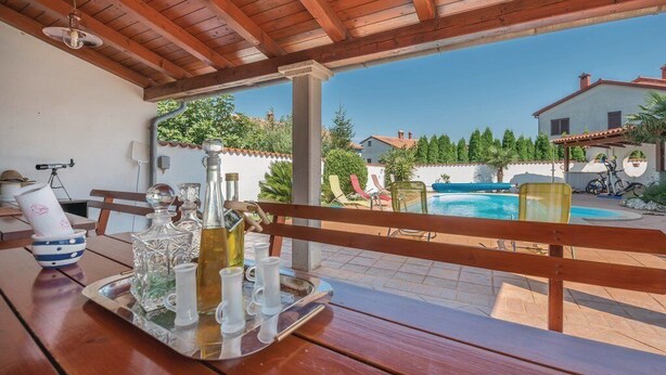A wooden table with a tray of glasses and a bottle, overlooking a swimming pool and a white building with a red roof.