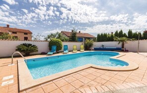 Ein Pool mit geschwungener Kante, umgeben von einer gefliesten Liegefläche. Mehrere Liegestühle und eine Palme sind vorhanden.