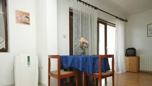 A dining area with a table covered by a blue tablecloth, two wooden chairs, a refrigerator, a television, and a window with white curtains.